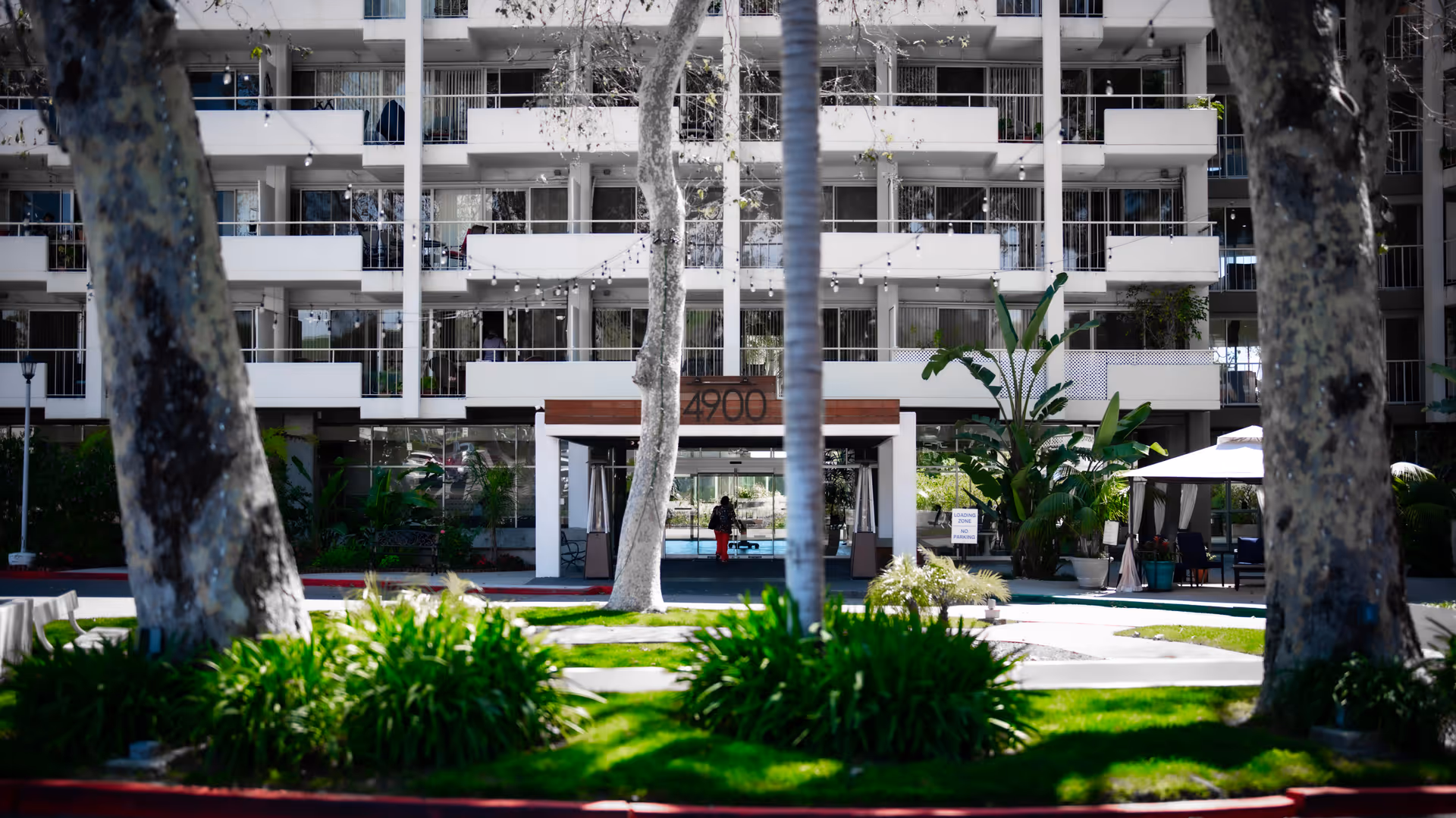Exterior view of a multi-story residential building with balconies, surrounded by trees and green landscaping. There is a covered entrance with the number 4900 above it, and outdoor seating areas with umbrellas visible on the right side.