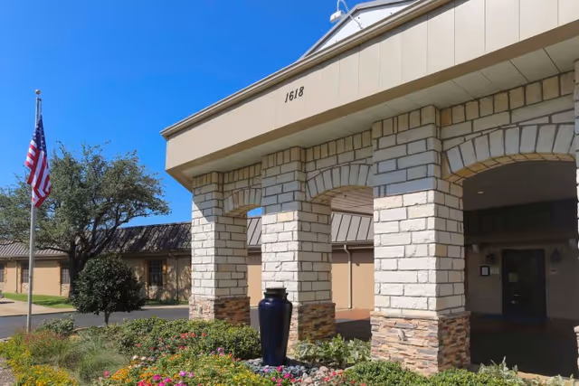 Exterior view of Carrollton Health & Rehabilitation Center showing the entrance with stone pillars, a covered driveway, a garden with flowers and a large blue vase, an American flag on a flagpole, and a clear blue sky.