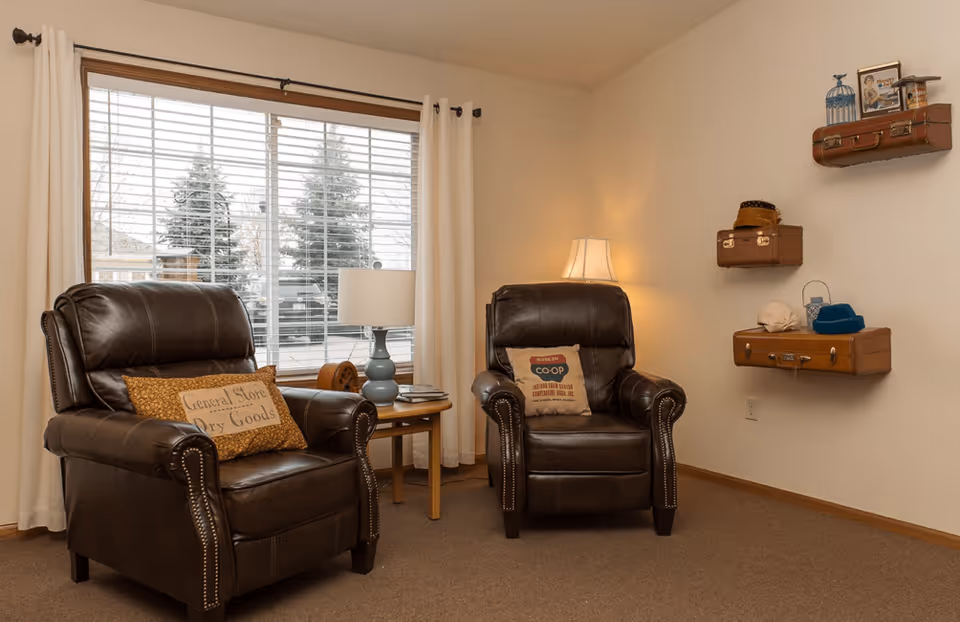 A cozy living room area with two dark brown leather armchairs, each with a decorative pillow. Between the chairs is a small wooden side table with a blue lamp and some books. Behind the chairs is a large window with white curtains and blinds, showing trees outside. On the right wall, there are three floating shelves made from vintage suitcases holding decorative items including hats and framed pictures. The room has beige walls and carpeted floor.