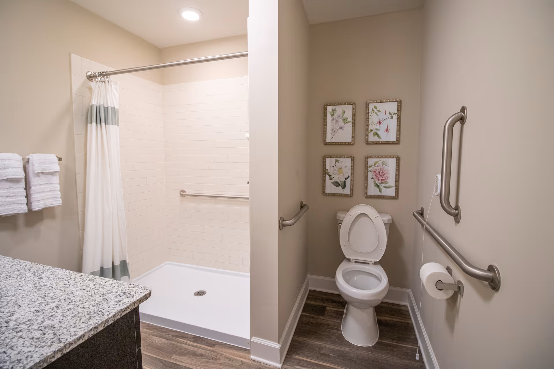 A clean and accessible bathroom featuring a walk-in shower with a white and gray striped curtain, grab bars on the walls, a toilet with the seat up, four framed floral pictures above the toilet, a granite countertop, and neatly folded white towels hanging on a towel rack.