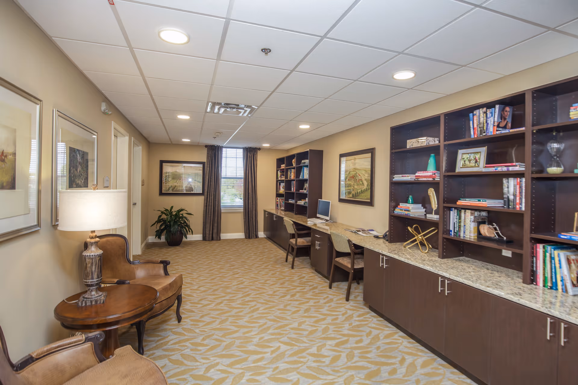 Long furnished common room with bookshelves, a desk/workstation area with computers, seating, and decorative artwork.