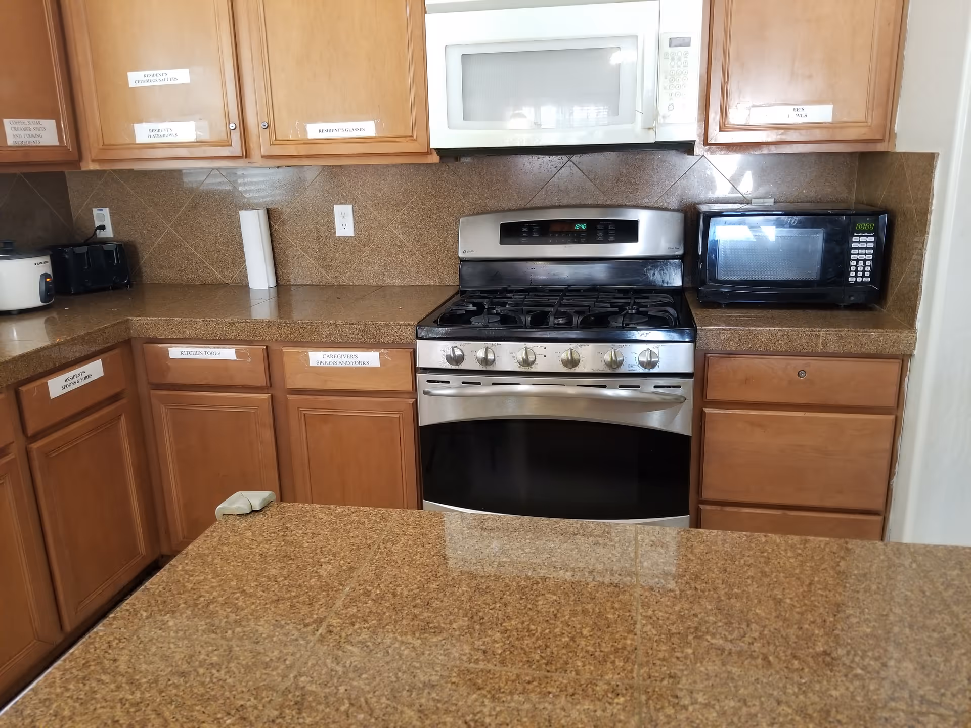 A kitchen area with brown granite countertops and wooden cabinets. The kitchen features a stainless steel gas stove with an oven, a white microwave above the stove, and a black microwave on the right countertop. Various cabinets and drawers have labels indicating their contents such as 'RESIDENT'S SPOONS & FORKS', 'KITCHEN TOOLS', 'CAREGIVERS SPOONS AND FORKS', and others. There is also a rice cooker and a toaster on the left countertop.