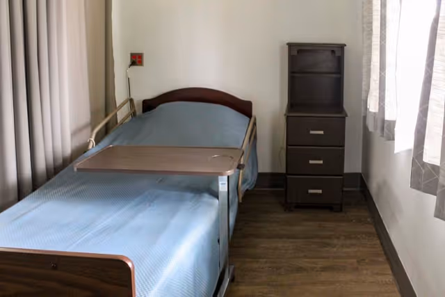 A single adjustable hospital-style bed with a blue blanket and overbed table beside a small three-drawer nightstand in a sparsely furnished room with curtains.