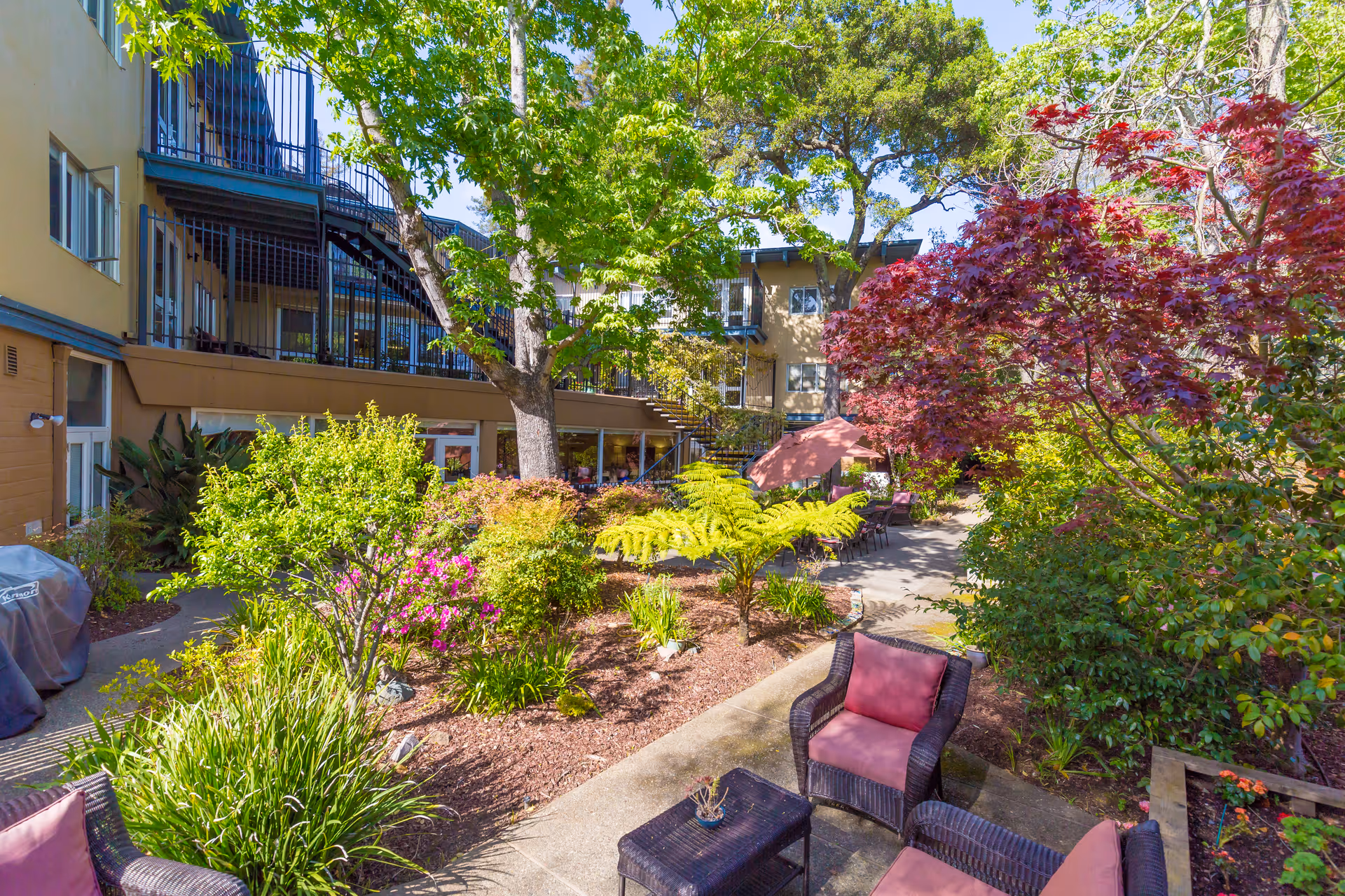 Sunlit landscaped courtyard with patio seating, umbrellas, and a surrounding multi-story memory care building.