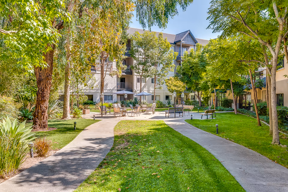 Sunlit landscaped courtyard with paved paths, benches, tables and trees in front of a multi-story senior living building.