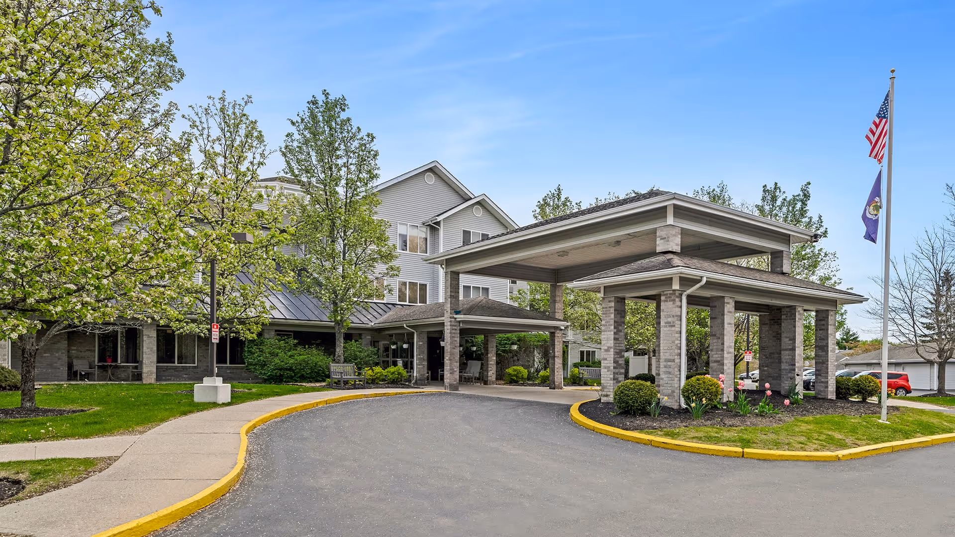 Exterior view of Holiday Woods at Canco senior living facility showing the main entrance with a covered drop-off area, surrounded by trees and landscaped greenery under a clear blue sky.