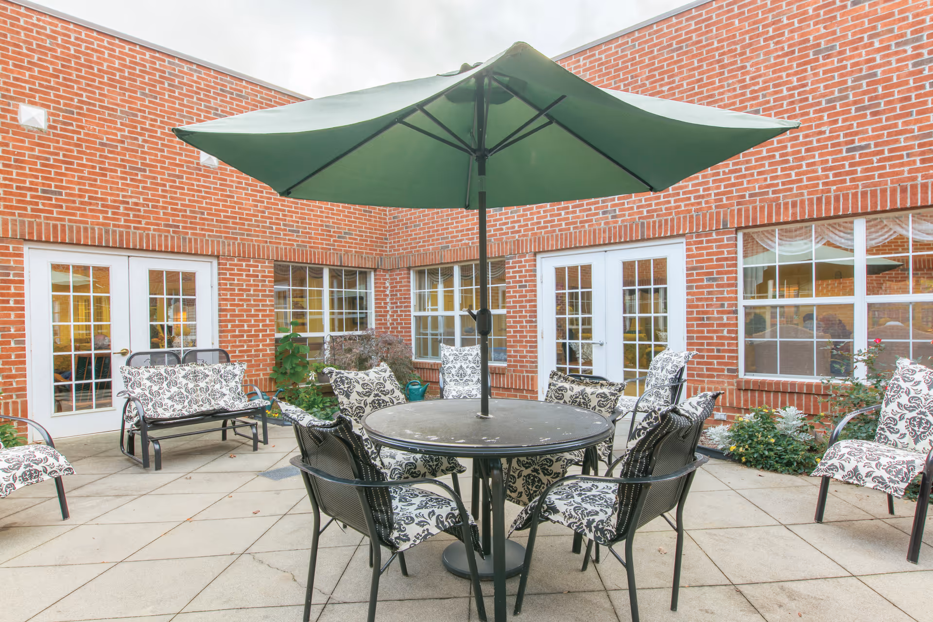 Outdoor brick courtyard patio with a round table, green umbrella, and patterned cushioned chairs in front of French doors and windows.