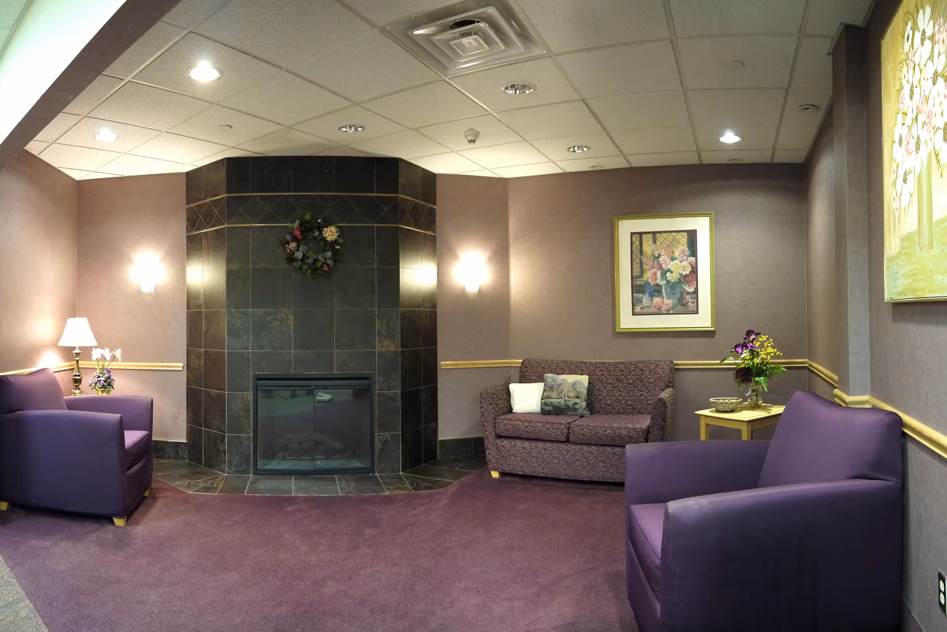 A cozy lounge area with purple carpeting and walls, featuring a dark tiled fireplace with a floral wreath above it. The room has two purple armchairs, a patterned loveseat with decorative pillows, and two side tables with lamps and flower arrangements. Framed floral artwork hangs on the walls.