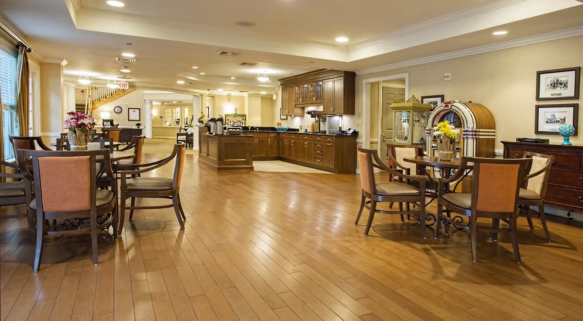 Spacious, well-lit communal dining and lounge area with wooden floors, round tables and chairs and a kitchenette/bar in the background.