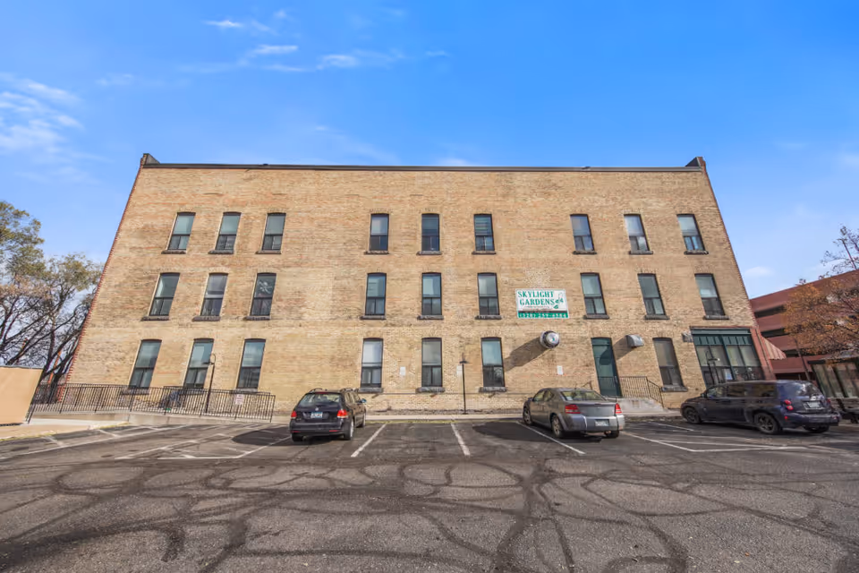Exterior view of a three-story brick building with multiple windows and a parking lot in front. There are a few parked cars and a sign on the building that reads 'Skylight Gardens'. The sky is clear and blue.