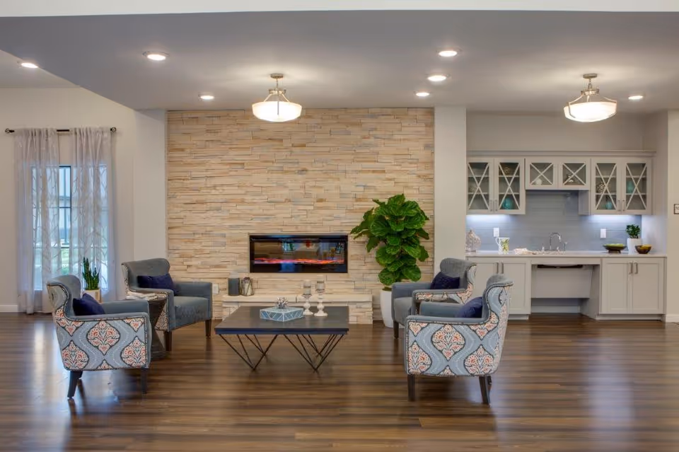 Communal living area with patterned armchairs around a coffee table facing a modern recessed fireplace and a small kitchenette.