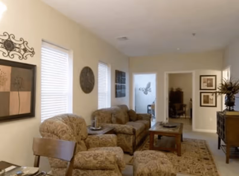 A cozy living room with beige walls and carpet, featuring a patterned sofa, armchair, and ottoman. There is a wooden coffee table in front of the sofa, wall art including framed pictures and decorative pieces, and two windows with blinds letting in natural light. The room leads to other areas of the home through open doorways.