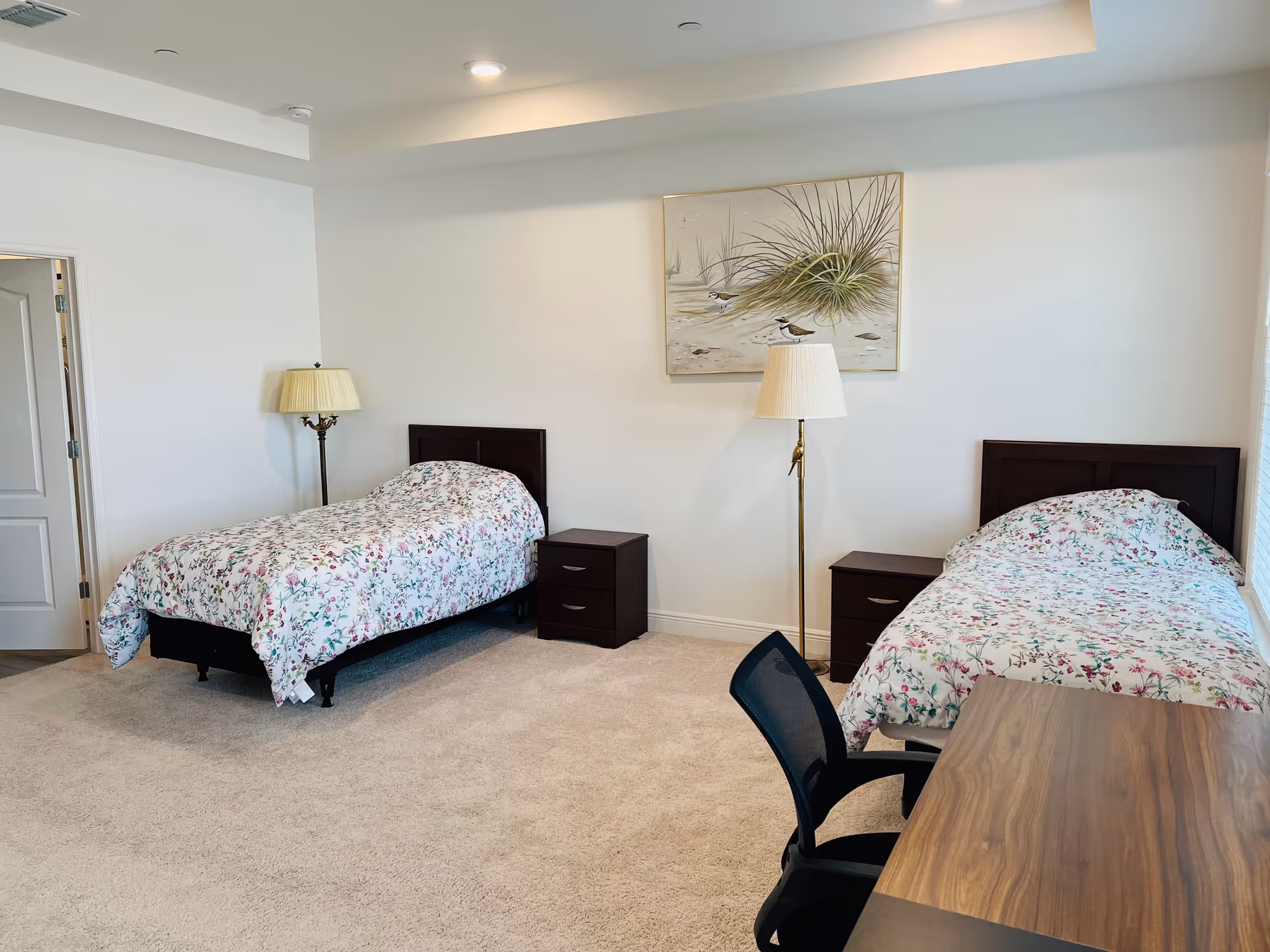 A bright bedroom with two single beds, each with floral bedspreads. Between the beds are two dark wooden nightstands, each with a lamp. A painting of birds and grass hangs on the wall above the right bed. There is a wooden desk with a black office chair in the foreground. The room has beige carpet and white walls.