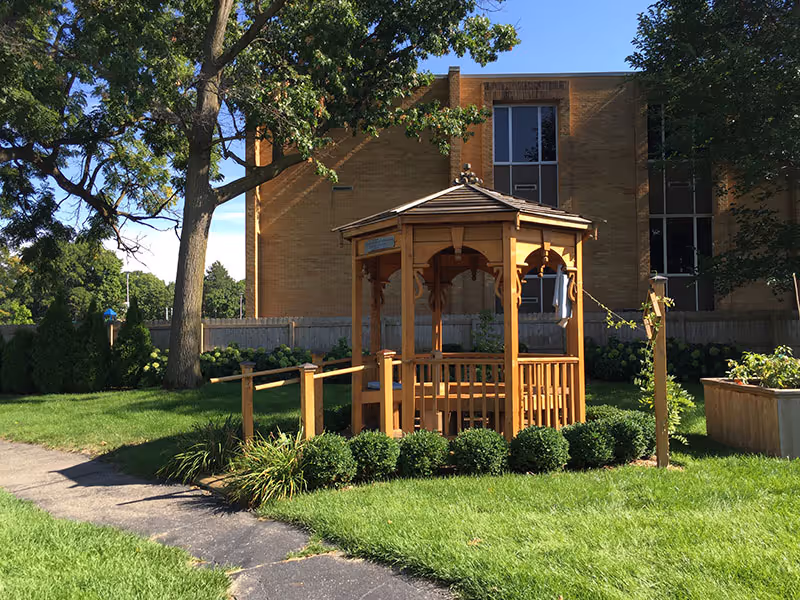 A wooden gazebo surrounded by green bushes and grass in an outdoor garden area with a large tree and a brick building in the background under a clear blue sky.