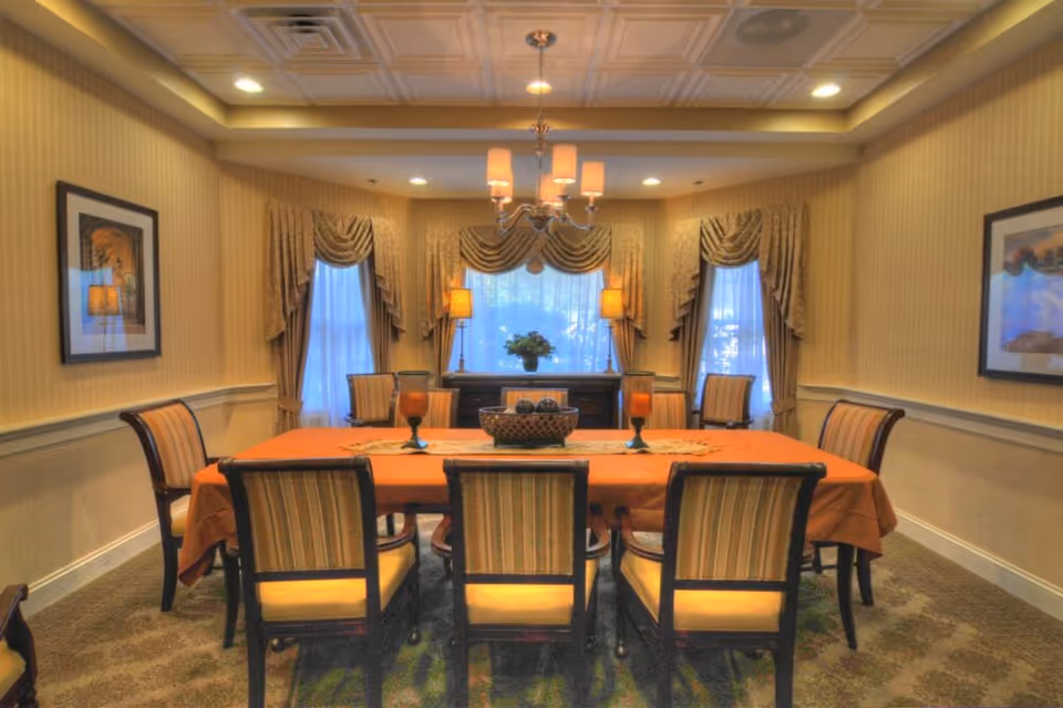 A formal dining room with a rectangular table covered with an orange tablecloth surrounded by eight striped upholstered chairs. The room has three windows with elegant draped curtains, two table lamps on a sideboard, a chandelier hanging from a coffered ceiling, and framed artwork on the walls.