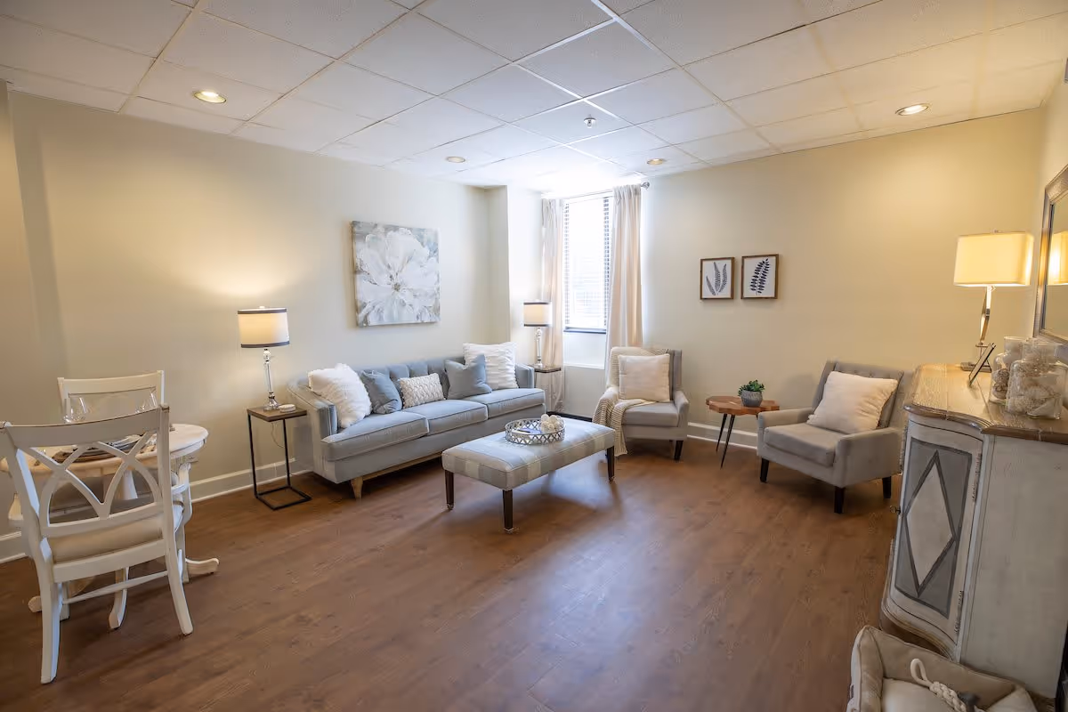 A cozy living room with a light gray sofa, two matching armchairs, a cushioned ottoman, and a small round wooden side table. The room has wooden flooring, soft lighting from table lamps, a window with white curtains, and decorative wall art including a large floral painting and two smaller framed prints.