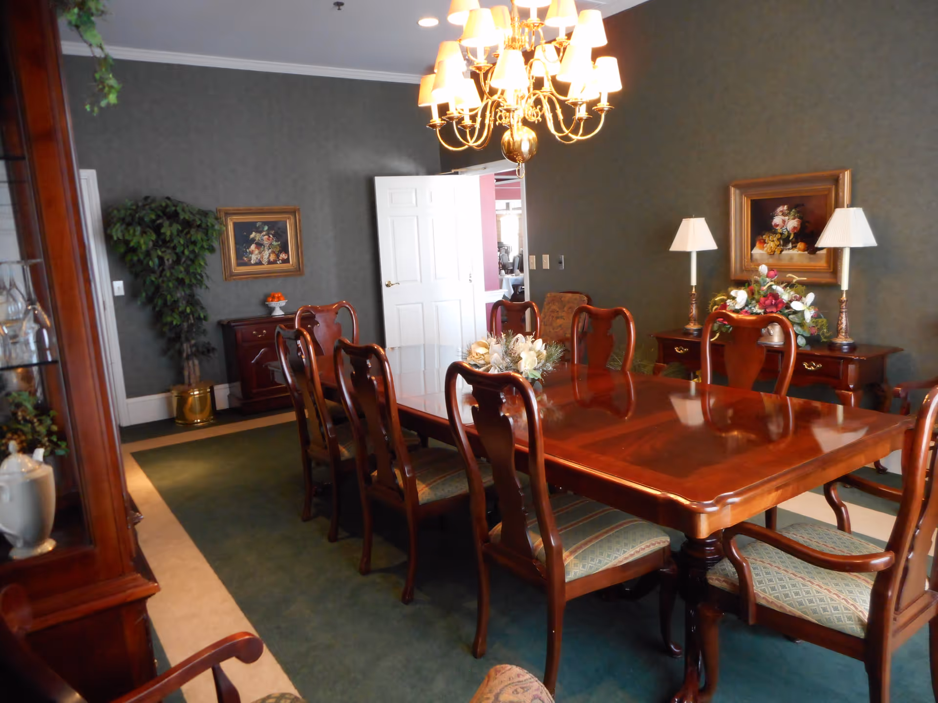 A formal dining room with a polished wooden dining table surrounded by eight matching wooden chairs with upholstered seats. The room features a green carpet with a beige border, dark green walls, two framed floral paintings, a chandelier with multiple lampshades hanging above the table, and two table lamps on a wooden sideboard. There is a potted plant in the corner and an open white door leading to another room.
