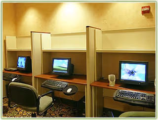 Three partitioned computer workstations with monitors and chairs in a beige-walled business center.