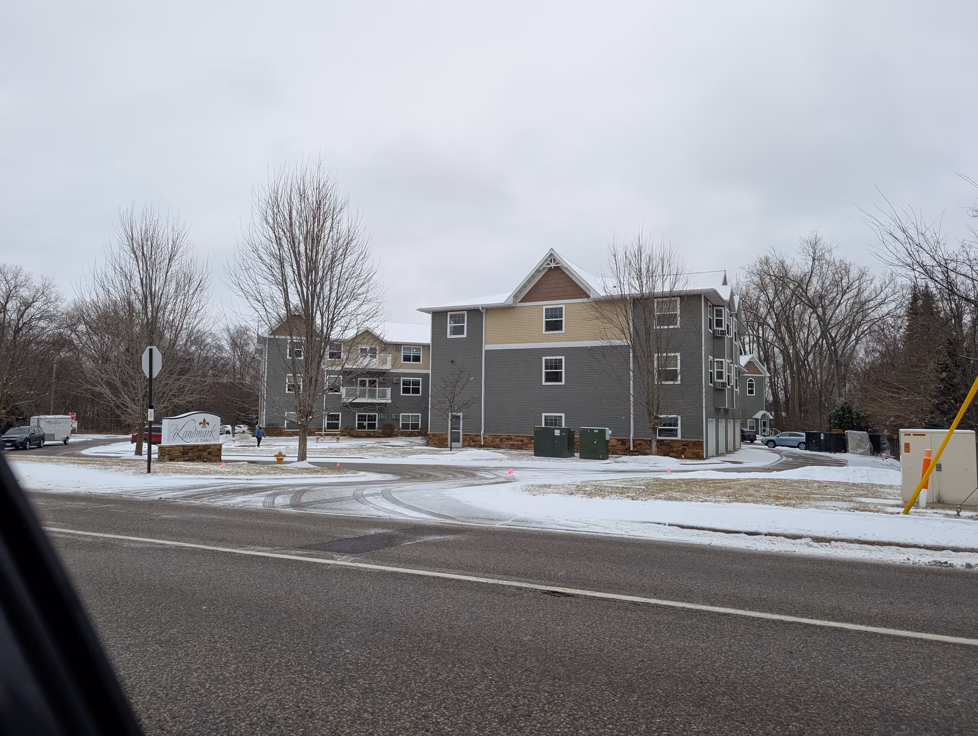 Exterior view of The Landmark Of Fridley building on a snowy day with leafless trees and a road in the foreground. The building is three stories tall with gray and beige siding and multiple windows. A sign with the facility name is visible near the entrance.