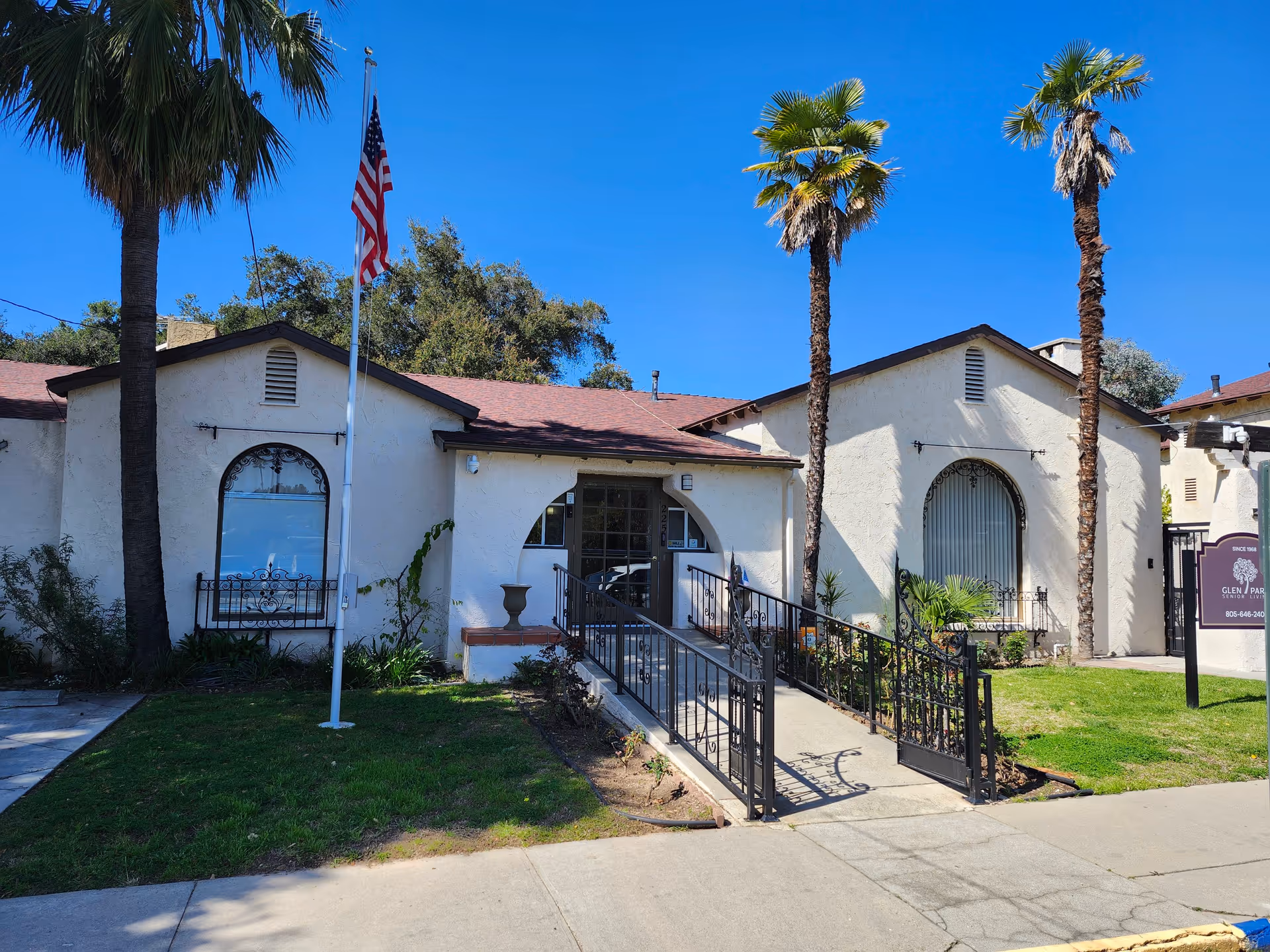 Exterior view of Glen Park at Ojai senior living facility showing a single-story building with a ramp entrance, two tall palm trees, an American flag on a flagpole, and a clear blue sky.