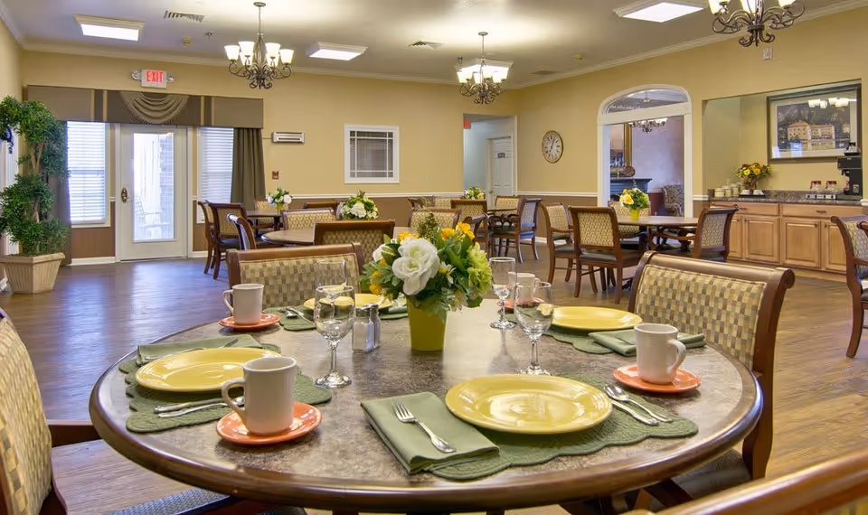 A bright and spacious dining room in a senior living facility with multiple round tables set with yellow plates, orange saucers, white mugs, green napkins, and silverware. Each table has a floral centerpiece. The room features wooden flooring, beige walls, chandeliers, and large windows with curtains allowing natural light. There is a door with an exit sign above it and a sideboard with cups and a coffee machine.