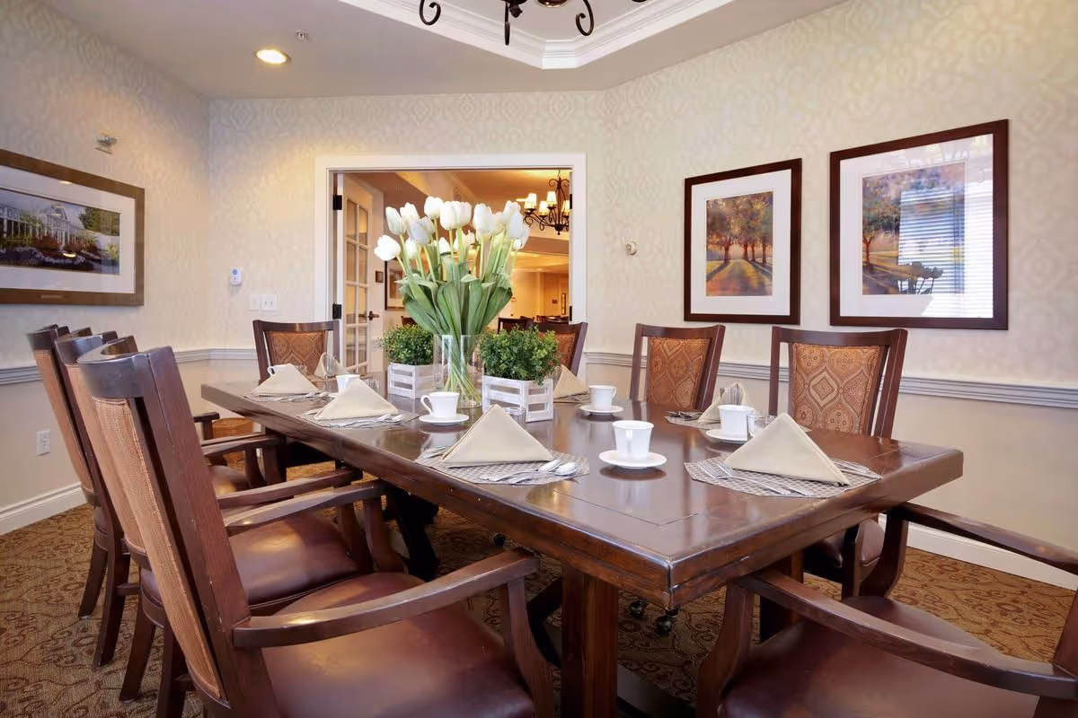 Spacious dining room with a long wooden table set with cups, folded napkins and a vase of white tulips, surrounded by wooden chairs and framed artwork.