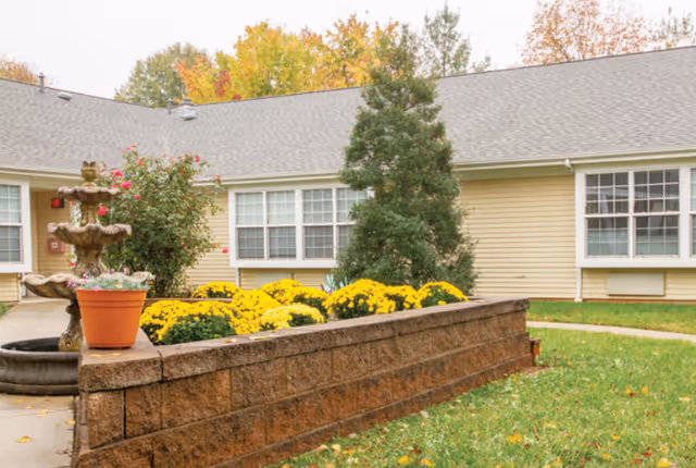 Courtyard in front of a single-story building with a low brick planter filled with yellow mums, a fountain, a potted plant, and windows.