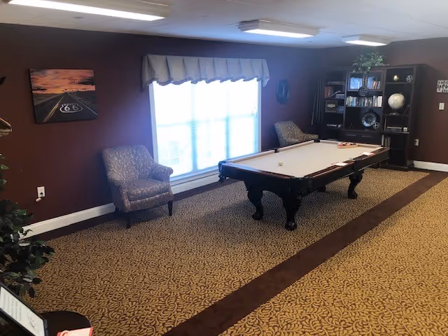 A room with a pool table in the center, two upholstered armchairs near a large window with blinds and a valance, a bookshelf with various items including books and a globe, and a patterned carpet covering the floor.