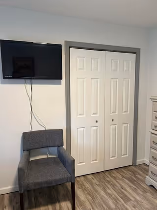 Small room interior with a wall-mounted TV above a gray upholstered chair beside white double closet doors and a dresser edge.