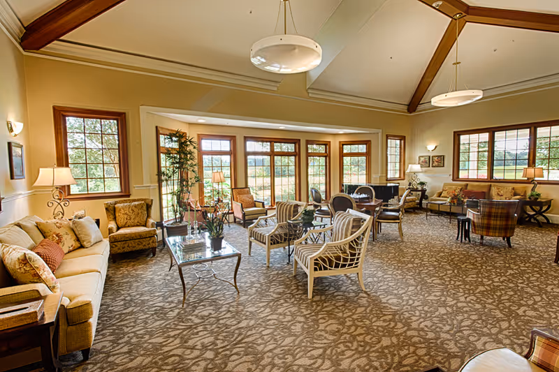 A spacious and well-lit living room with large windows allowing natural light to fill the space. The room features multiple seating areas including sofas, armchairs, and small tables arranged on a patterned carpet. The ceiling has exposed wooden beams and modern hanging light fixtures. The decor includes plants, lamps, and framed artwork on the walls, creating a warm and inviting atmosphere.