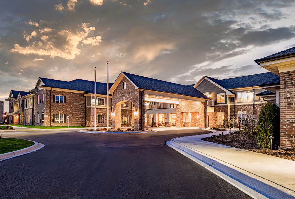 Front exterior of a brick senior living facility with an illuminated covered entrance and circular driveway at dusk.