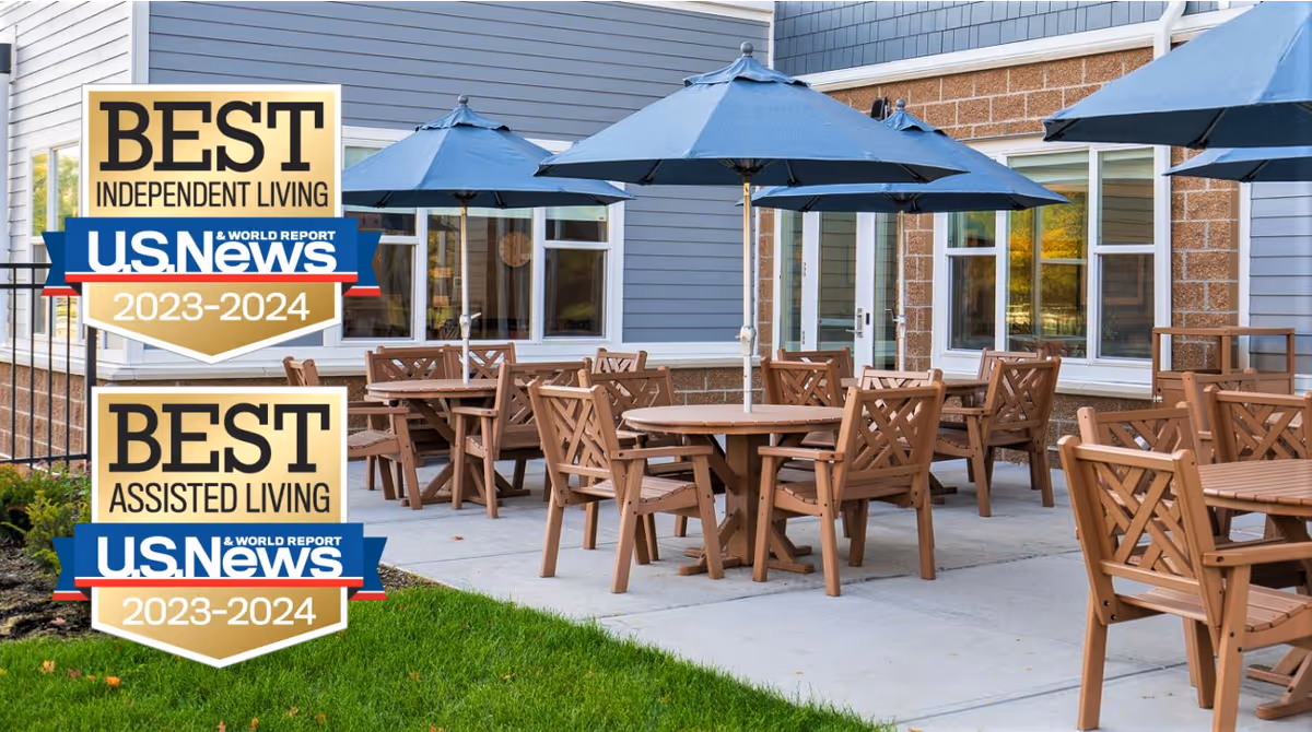 Outdoor patio area at Willows Bend Senior Living with multiple wooden tables and chairs under blue umbrellas, adjacent to a building with gray siding and large windows.