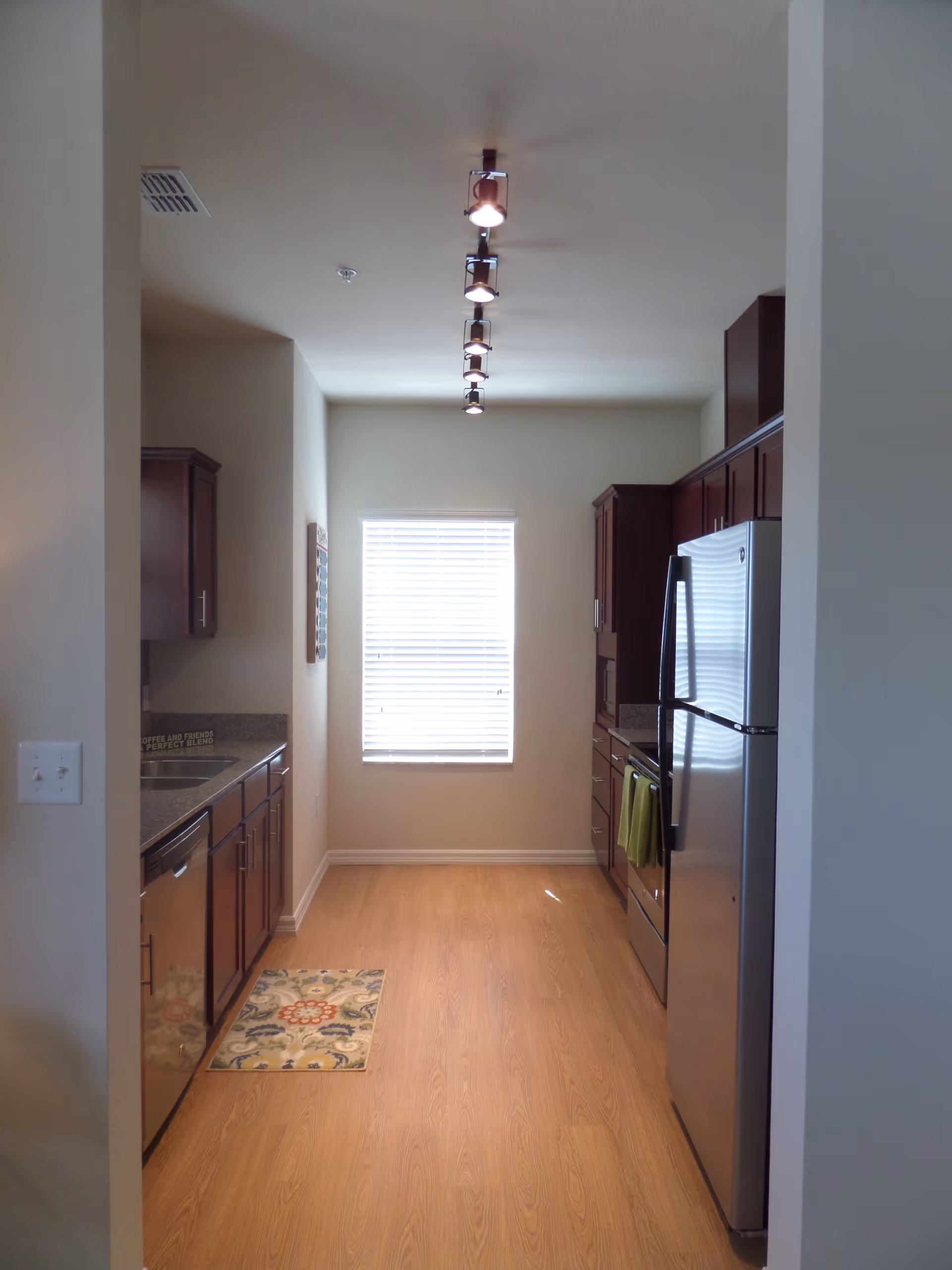 A narrow kitchen with wooden cabinets on both sides, a stainless steel refrigerator on the right, a dishwasher on the left, a window with blinds at the end, and a track lighting fixture on the ceiling. The floor is wooden, and there is a small patterned rug near the left side.