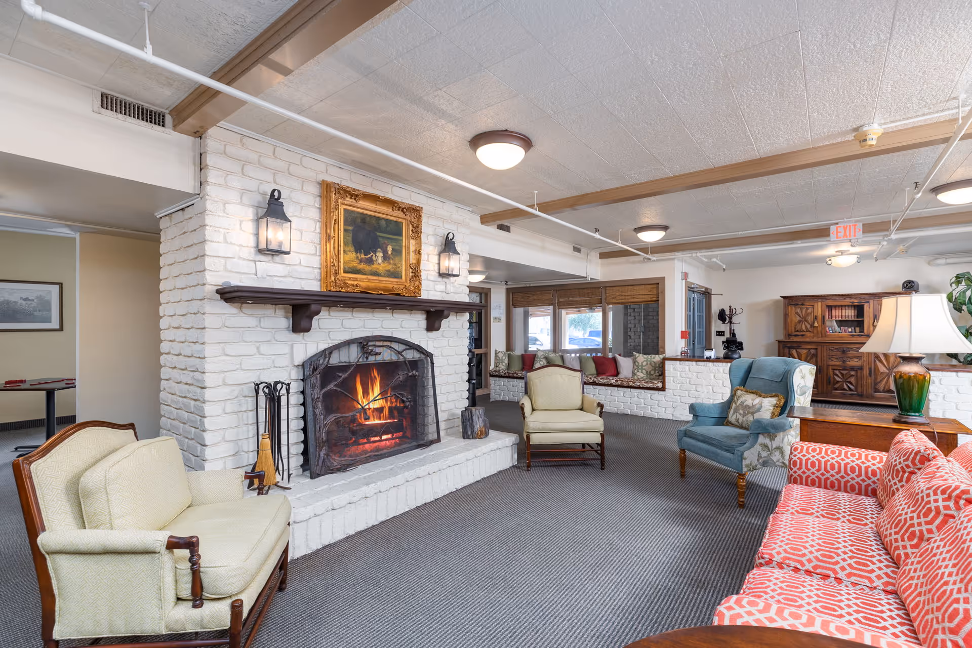 A cozy living room area with a lit fireplace set in a white brick wall. The room features various seating options including a light green loveseat, a blue armchair, a red patterned sofa, and a beige armchair. Above the fireplace is a framed painting of cows. The room has carpeted flooring, ceiling lights, and a window seat with cushions near large windows.
