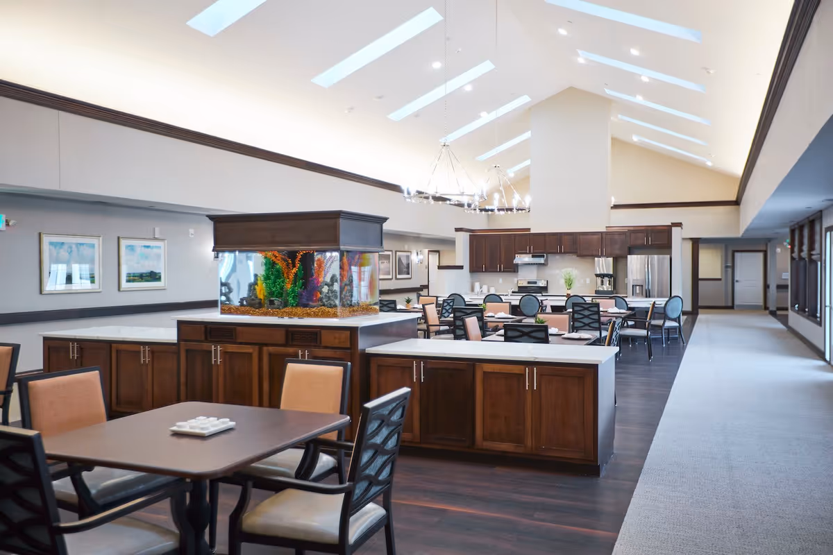 Bright communal dining and kitchen area with a central aquarium, multiple tables and chairs, and a vaulted skylight ceiling.