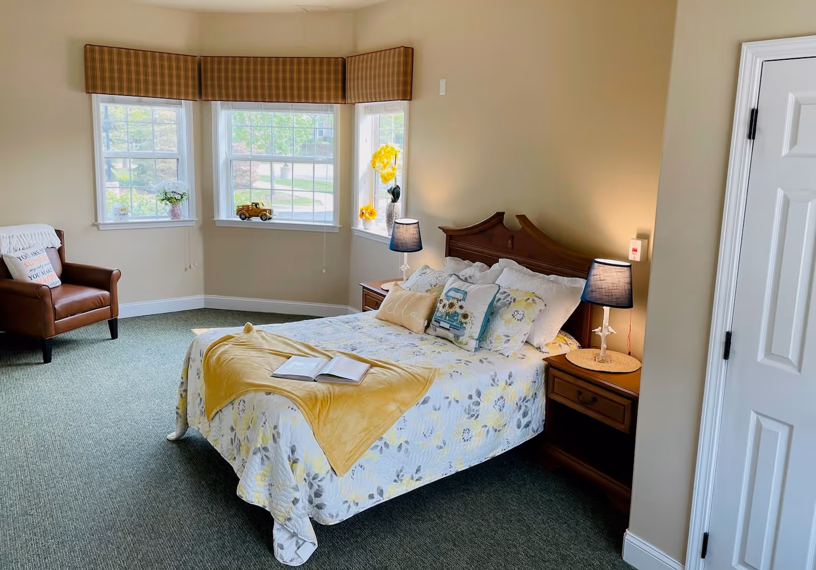 A cozy bedroom with a bed covered in floral bedding and a yellow throw blanket, two bedside tables with lamps, a brown armchair with a pillow, and three windows with plaid valances letting in natural light.