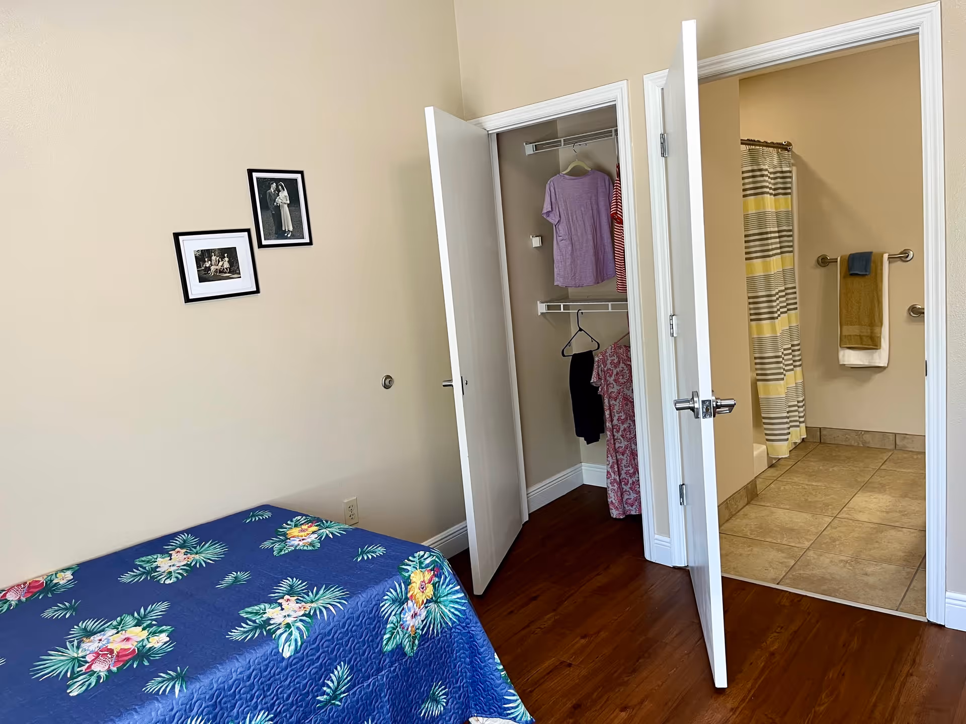 A bedroom with a bed covered in a blue floral bedspread, two framed black and white photos on a beige wall, an open closet with clothes hanging inside, and an open door leading to a bathroom with a striped shower curtain and towels hanging on a rack.