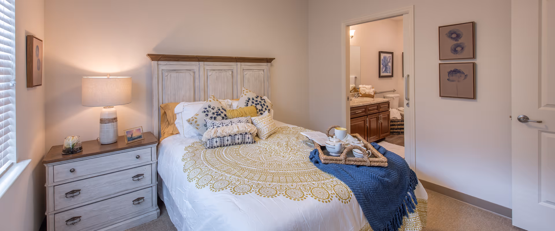 Cozy bedroom with a decorated bed, nightstand and lamp, and an open door leading to an en-suite bathroom.