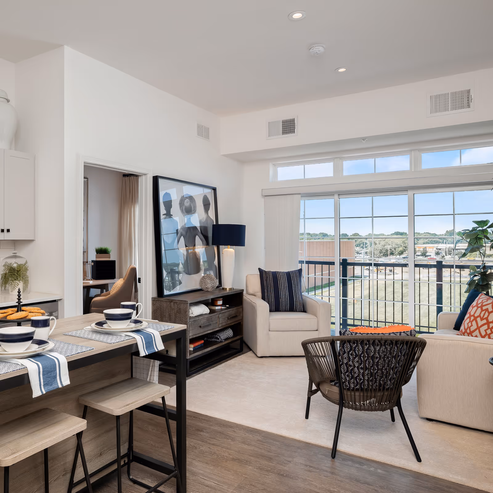 Bright, modern open-plan living room with a breakfast bar, armchairs, a side table with lamp, and large windows/sliding door to a balcony.