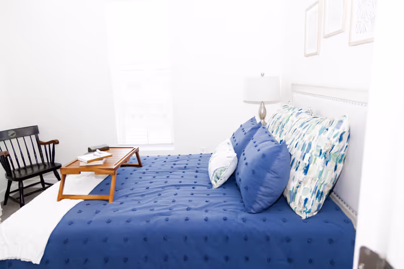 A bright bedroom with a bed covered in a blue textured comforter and multiple pillows with blue and patterned pillowcases. A wooden tray with books and glasses sits on the bed. There is a black wooden chair with armrests next to the bed and a window with blinds letting in natural light. A lamp with a white shade is on a bedside table, and framed artwork hangs on the wall above the bed.