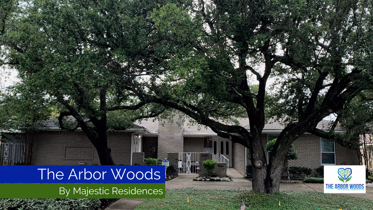 Front exterior view of a single-story brick building partially obscured by large leafy trees, with a walkway leading to a double-door entrance. There is a sign in the bottom right corner with the logo and name 'The Arbor Woods by Majestic Residences.'
