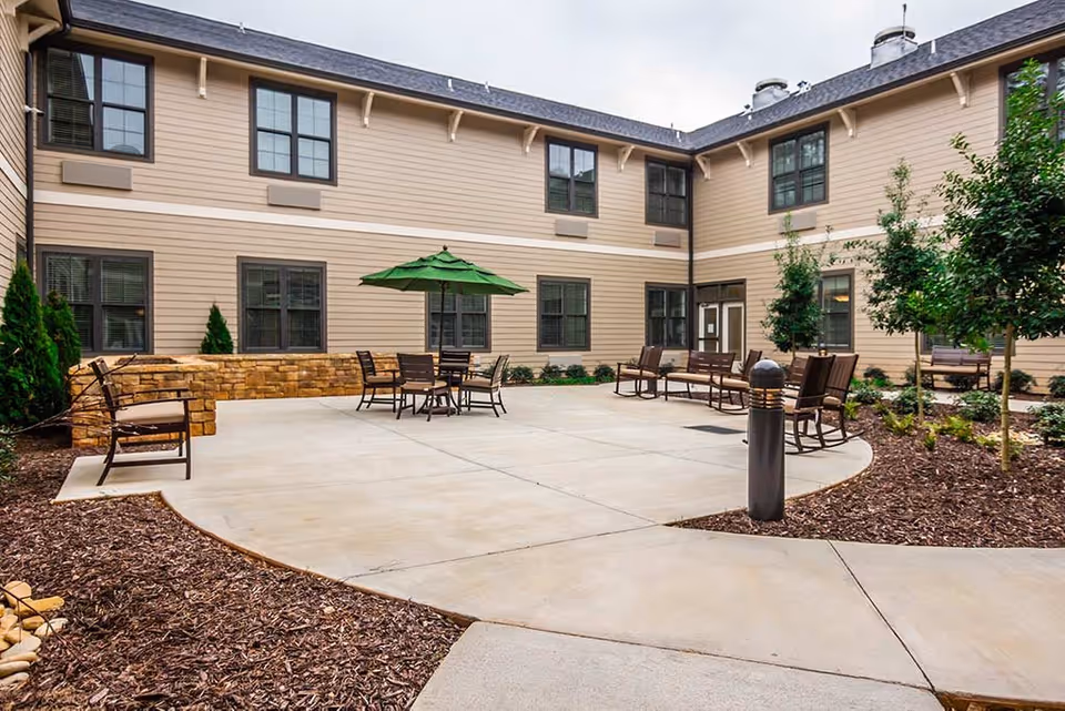 Outdoor courtyard area of a senior living facility with beige two-story building walls, multiple windows, and a concrete patio. The patio has several seating arrangements including chairs, benches, and a table with a green umbrella. There are small trees and landscaped mulch beds surrounding the patio.