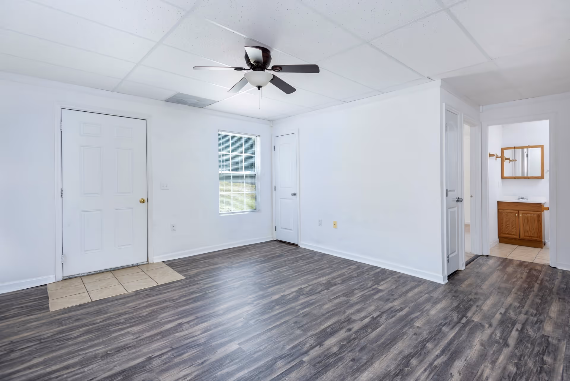 Empty bright living room with gray wood-look flooring, a ceiling fan, white walls, a window, and doors including one opening to a bathroom vanity.