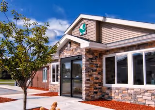 Front entrance of a single-story assisted living building with stone and siding facade, glass double doors, large windows, and landscaped walkways.