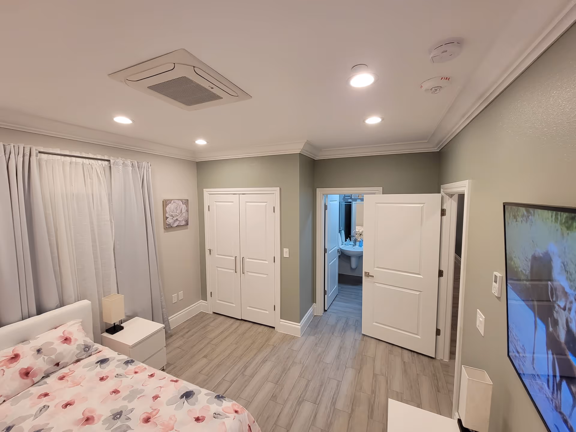 A clean and well-lit bedroom in an assisted living facility with a bed covered in floral bedding, a white nightstand with a lamp, a window with white curtains, a closet with double doors, an open door leading to a bathroom, and a wall-mounted TV showing an image of a moose.