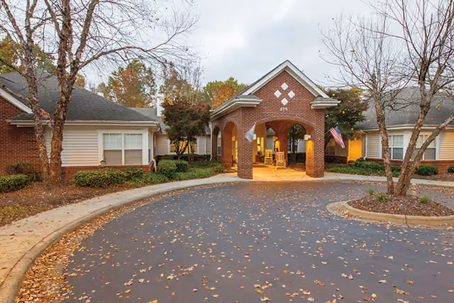 Exterior view of Brookdale Winston-Salem senior living facility showing a brick entrance with an arched portico, rocking chairs under the covered area, and surrounding single-story buildings with trees and autumn leaves on the ground.