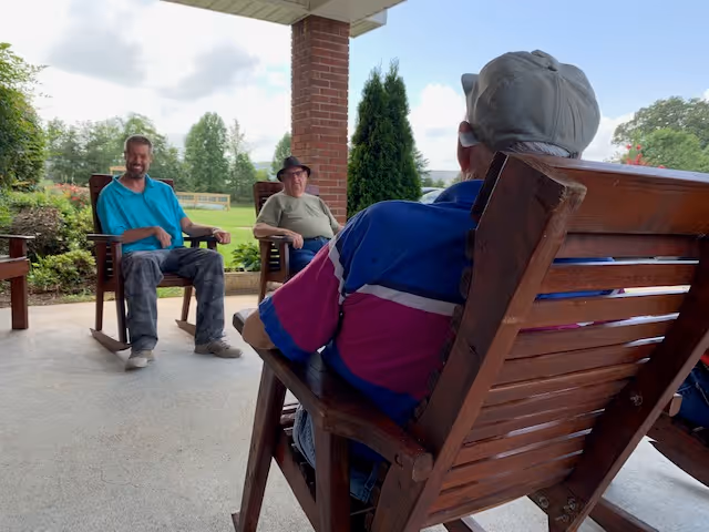 Three men sitting and relaxing on wooden rocking chairs on a covered patio with greenery and trees in the background under a partly cloudy sky.
