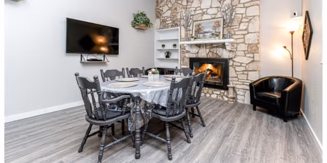 A cozy living room area with a round dining table covered with a white tablecloth and six black wooden chairs around it. There is a stone fireplace with a fire burning, a painting above the mantel, and a white shelving unit with plants and decor. A flat-screen TV is mounted on the wall to the left, and a black leather armchair is positioned near a tall floor lamp on the right. The room has light gray wood flooring and white walls.