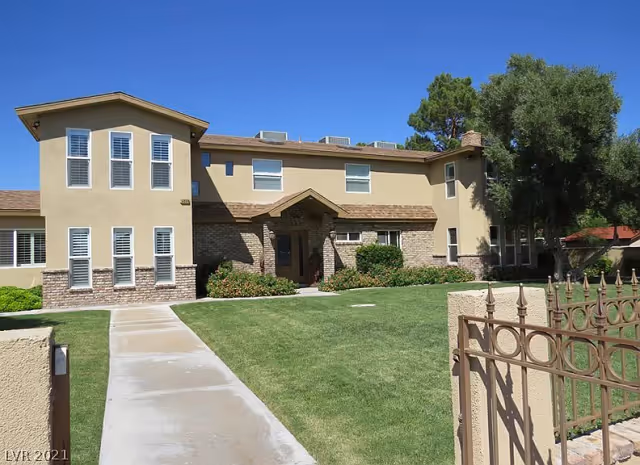 Exterior view of a two-story senior living facility building with beige walls, multiple windows with white shutters, a brown shingled roof, and a well-maintained green lawn with a concrete walkway leading to the entrance. There is a decorative metal fence and gate in the foreground, and trees are visible on the right side under a clear blue sky.