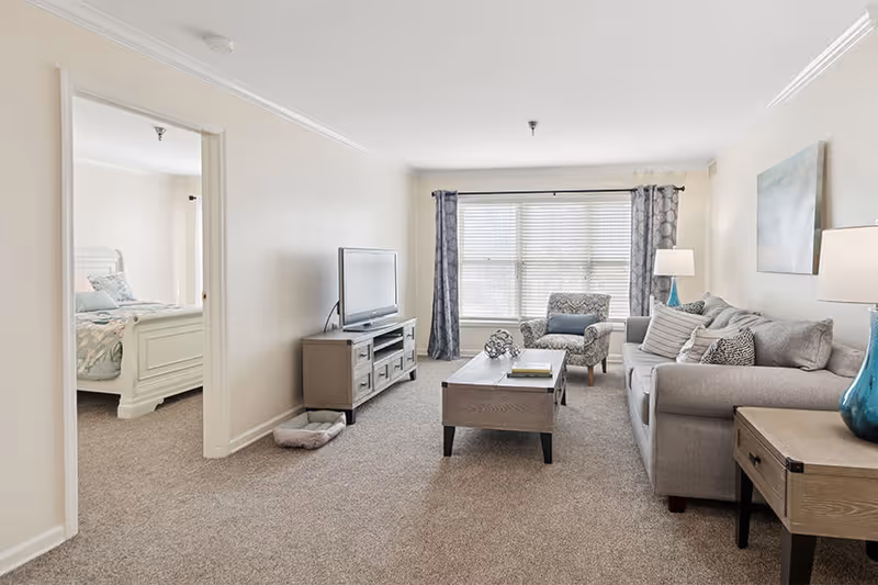 A bright living room with beige carpet and light-colored walls. The room features a gray sofa with multiple cushions, a patterned armchair, a wooden coffee table with decorative items and books, a TV on a wooden stand, and a side table with a blue lamp. Large windows with blinds and patterned curtains allow natural light to fill the space. An open doorway leads to a bedroom with a white bed frame and bedding.